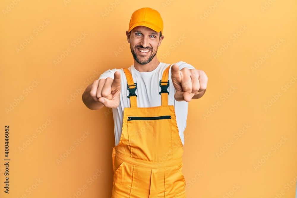 Young handsome man wearing handyman uniform over yellow background pointing to you and the camera with fingers, smiling positive and cheerful