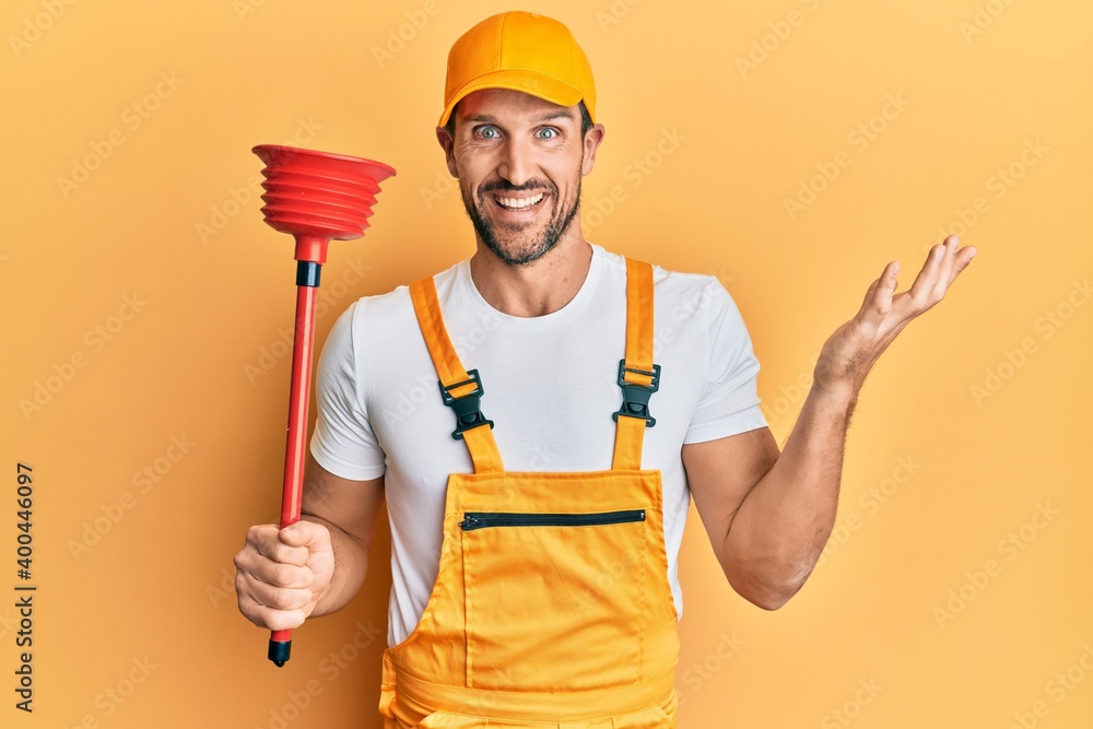 Young handsome man wearing plumber uniform holding toilet plunger celebrating achievement with happy smile and winner expression with raised hand Young handsome man wearing plumber uniform holding toilet plunger celebrating achievement with happy smile and winner expression with raised hand