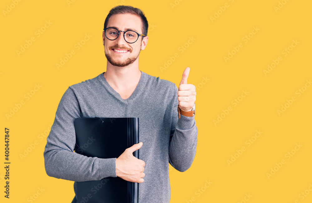 Young handsome caucasian man holding business folder smiling happy and positive, thumb up doing excellent and approval sign