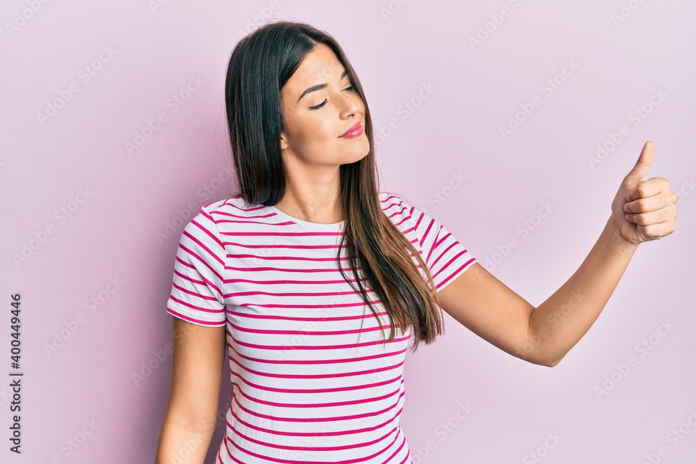 Young brunette woman wearing casual clothes over pink background looking proud, smiling doing thumbs up gesture to the side Young brunette woman wearing casual clothes over pink background looking proud, smiling doing thumbs up gesture to the side