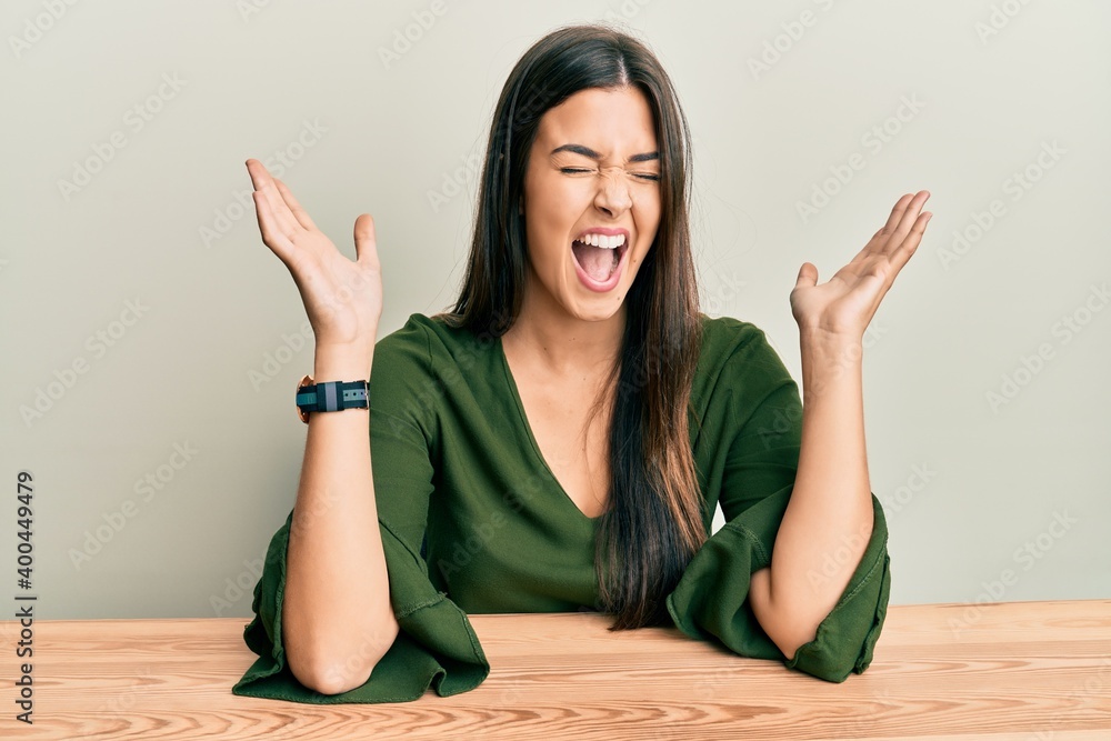 Young brunette woman wearing casual clothes sitting on the table celebrating mad and crazy for success with arms raised and closed eyes screaming excited. winner concept Young brunette woman wearing casual clothes sitting on the table celebrating mad and crazy for success with arms raised and closed eyes screaming excited. winner concept