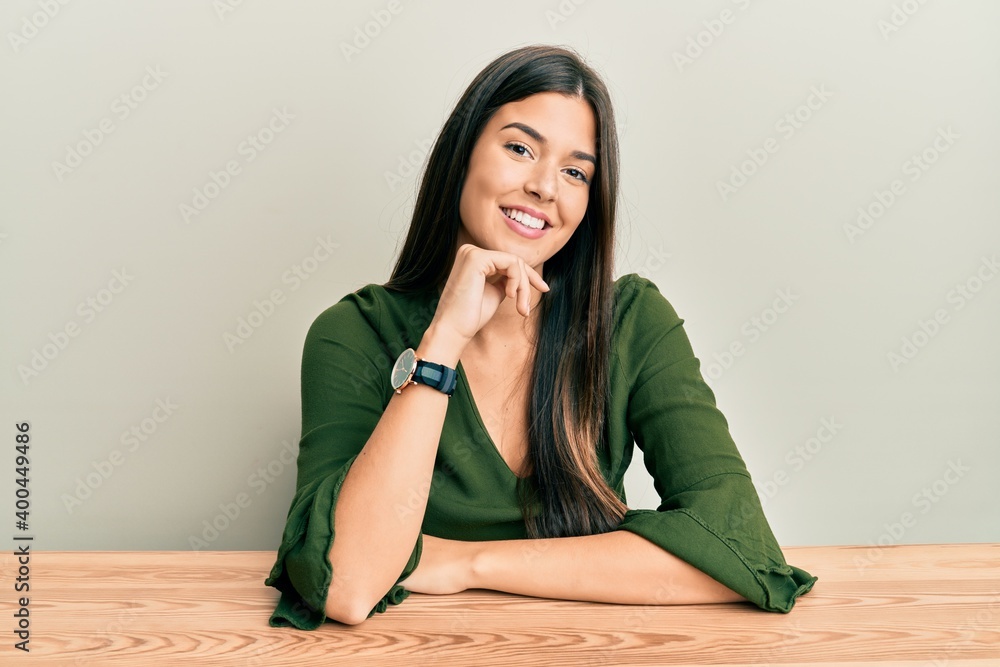 Young brunette woman wearing casual clothes sitting on the table looking confident at the camera with smile with crossed arms and hand raised on chin. thinking positive. Young brunette woman wearing casual clothes sitting on the table looking confident at the camera with smile with crossed arms and hand raised on chin. thinking positive.