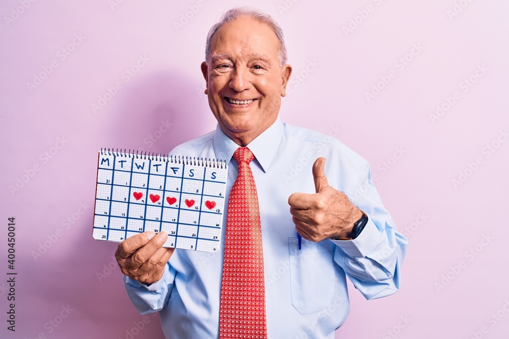 Senior handsome grey-haired businessman wearing tie holding cardio heart calendar smiling happy and positive, thumb up doing excellent and approval sign Senior handsome grey-haired businessman wearing tie holding cardio heart calendar smiling happy and positive, thumb up doing excellent and approval sign