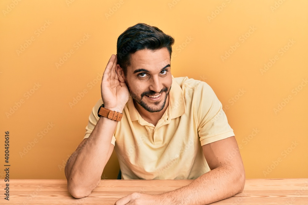 Young hispanic man wearing casual clothes sitting on the table smiling with hand over ear listening an hearing to rumor or gossip. deafness concept. Young hispanic man wearing casual clothes sitting on the table smiling with hand over ear listening an hearing to rumor or gossip. deafness concept.