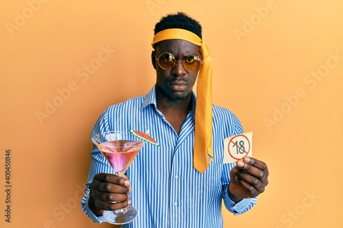 Handsome black man drunk wearing tie over head drinking cocktail holding under 18 sign clueless and confused expression. doubt concept.