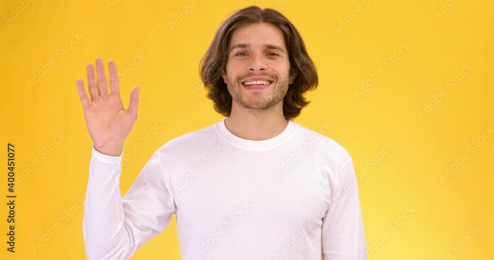 Studio shot of young friendly talkative guy waving hand and saying Hello, slow motion, orange background