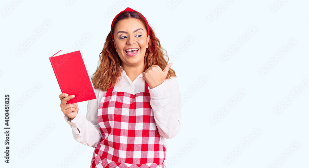 Young latin woman wearing apron holding recipe book pointing thumb up to the side smiling happy with open mouth Young latin woman wearing apron holding recipe book pointing thumb up to the side smiling happy with open mouth