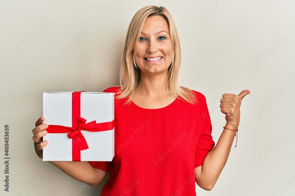 Beautiful middle age blonde woman holding gift smiling happy and positive, thumb up doing excellent and approval sign Beautiful middle age blonde woman holding gift smiling happy and positive, thumb up doing excellent and approval sign