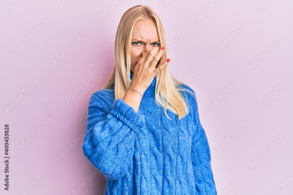 Young blonde girl wearing wool winter sweater smelling something stinky and disgusting, intolerable smell, holding breath with fingers on nose. bad smell Young blonde girl wearing wool winter sweater smelling something stinky and disgusting, intolerable smell, holding breath with fingers on nose. bad smell