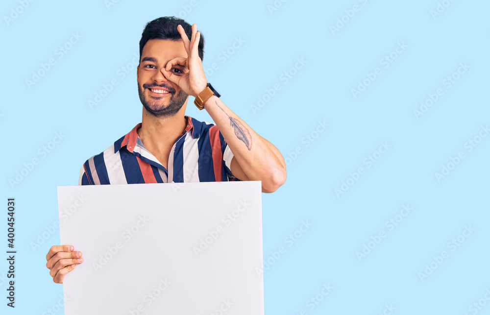 Young handsome hispanic man holding cardboard banner with blank space smiling happy doing ok sign with hand on eye looking through fingers Young handsome hispanic man holding cardboard banner with blank space smiling happy doing ok sign with hand on eye looking through fingers