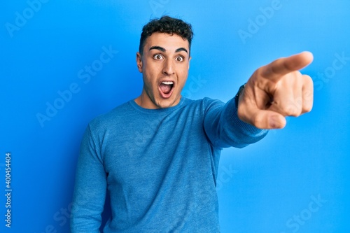 Hispanic young man wearing casual winter sweater pointing with finger surprised ahead, open mouth amazed expression, something on the front