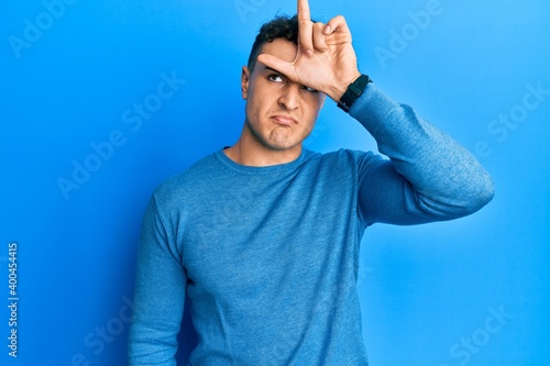 Hispanic young man wearing casual winter sweater making fun of people with fingers on forehead doing loser gesture mocking and insulting.