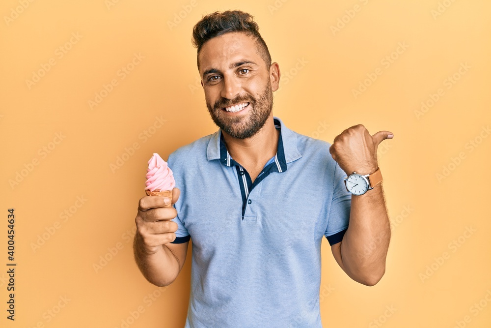 Handsome man with beard eating ice cream cone pointing thumb up to the side smiling happy with open mouth Handsome man with beard eating ice cream cone pointing thumb up to the side smiling happy with open mouth