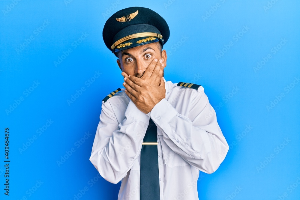 Handsome man with beard wearing airplane pilot uniform shocked covering mouth with hands for mistake. secret concept. Handsome man with beard wearing airplane pilot uniform shocked covering mouth with hands for mistake. secret concept.