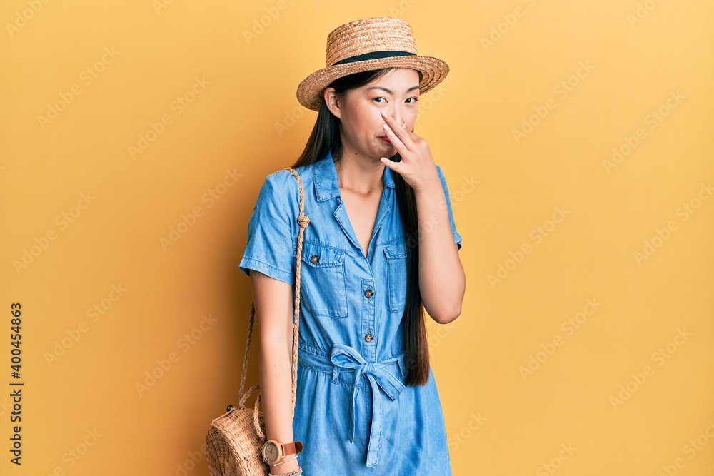 Young chinese woman wearing summer hat smelling something stinky and disgusting, intolerable smell, holding breath with fingers on nose. bad smell