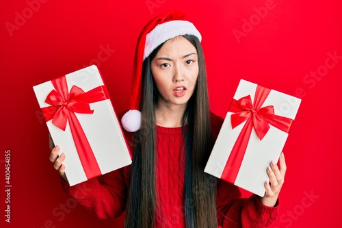 Young chinese woman wearing christmas hat and holding gifts in shock face, looking skeptical and sarcastic, surprised with open mouth