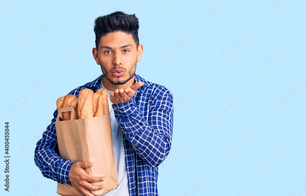 Handsome latin american young man holding paper bag with bread looking at the camera blowing a kiss with hand on air being lovely and sexy. love expression. Handsome latin american young man holding paper bag with bread looking at the camera blowing a kiss with hand on air being lovely and sexy. love expression.