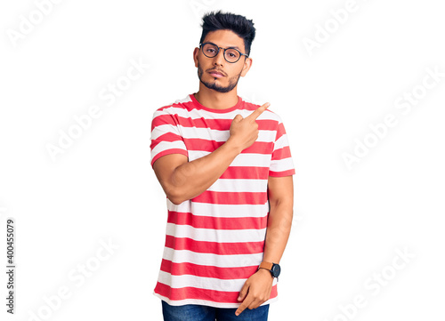 Handsome latin american young man wearing casual clothes and glasses pointing with hand finger to the side showing advertisement, serious and calm face