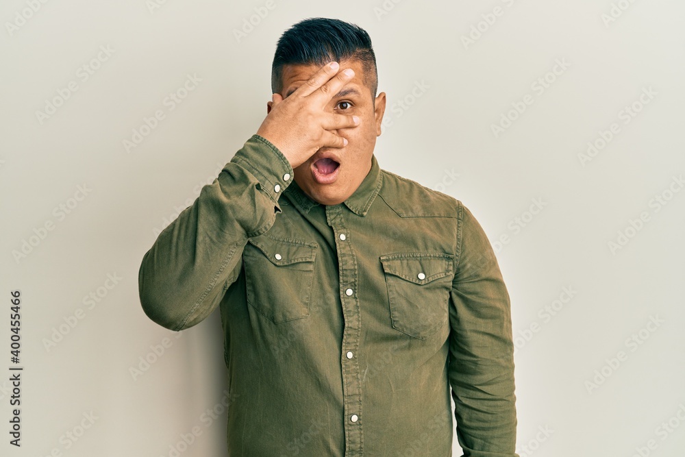 Young latin man wearing casual clothes peeking in shock covering face and eyes with hand, looking through fingers with embarrassed expression. Young latin man wearing casual clothes peeking in shock covering face and eyes with hand, looking through fingers with embarrassed expression.