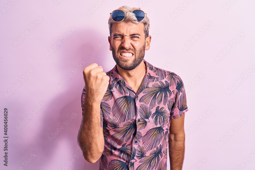 Naklejka premium Handsome blond man on vacation wearing casual shirt and sunglasses over pink background angry and mad raising fist frustrated and furious while shouting with anger. Rage and aggressive concept.