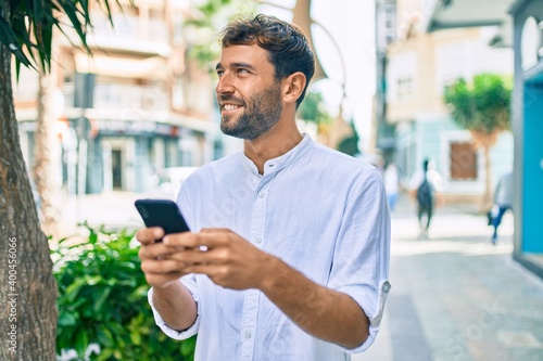 Handsome man with beard wearing casual white shirt on a sunny day smiling happy outdoors using smartphone