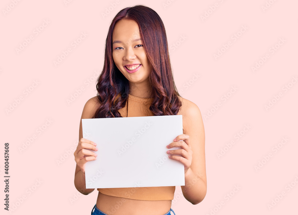 Young beautiful chinese girl holding paper banner with blank space looking positive and happy standing and smiling with a confident smile showing teeth Young beautiful chinese girl holding paper banner with blank space looking positive and happy standing and smiling with a confident smile showing teeth