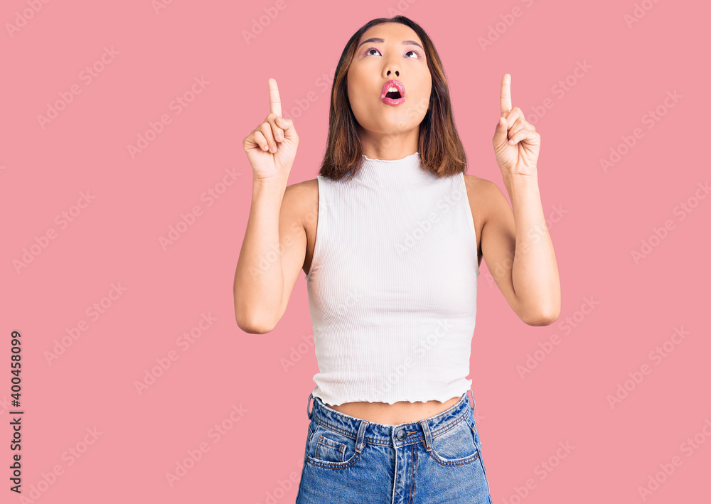 Young beautiful chinese girl wearing casual clothes amazed and surprised looking up and pointing with fingers and raised arms. Young beautiful chinese girl wearing casual clothes amazed and surprised looking up and pointing with fingers and raised arms.