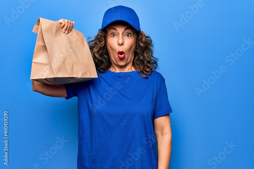 Middle age beautiful delivery woman holding deliver paper bag with takeaway food scared and amazed with open mouth for surprise, disbelief face