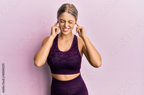 Beautiful blonde woman wearing sportswear over pink background covering ears with fingers with annoyed expression for the noise of loud music. deaf concept.