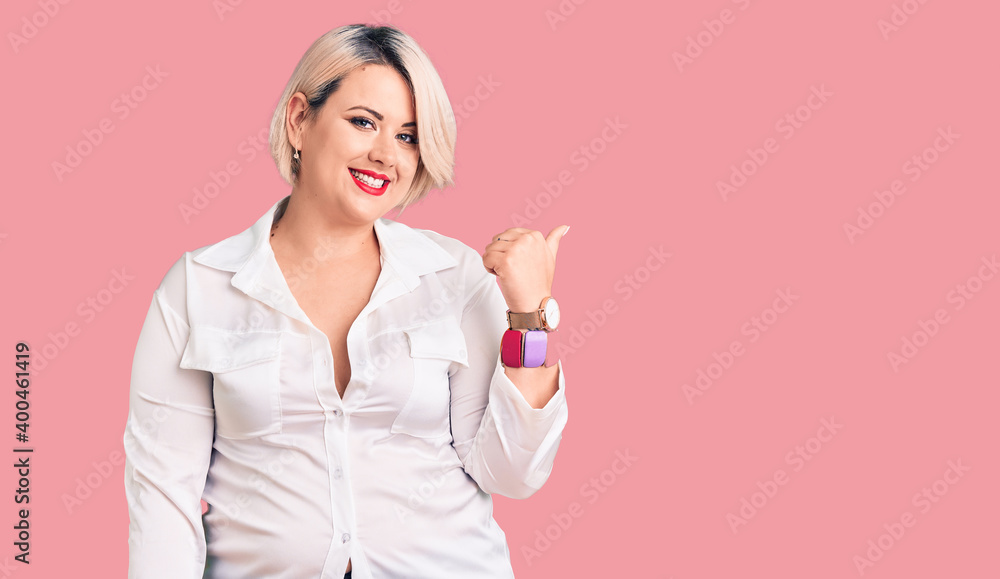 Young blonde plus size woman wearing casual shirt smiling with happy face looking and pointing to the side with thumb up. Young blonde plus size woman wearing casual shirt smiling with happy face looking and pointing to the side with thumb up.