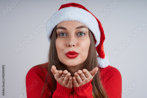 Close-up portrait of beautiful young woman in red sweater and santa hat blowing a kiss  on gray background.