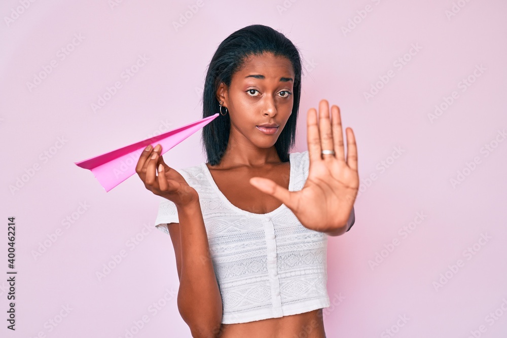 Young african american woman holding paper airplane with open hand doing stop sign with serious and confident expression, defense gesture Young african american woman holding paper airplane with open hand doing stop sign with serious and confident expression, defense gesture