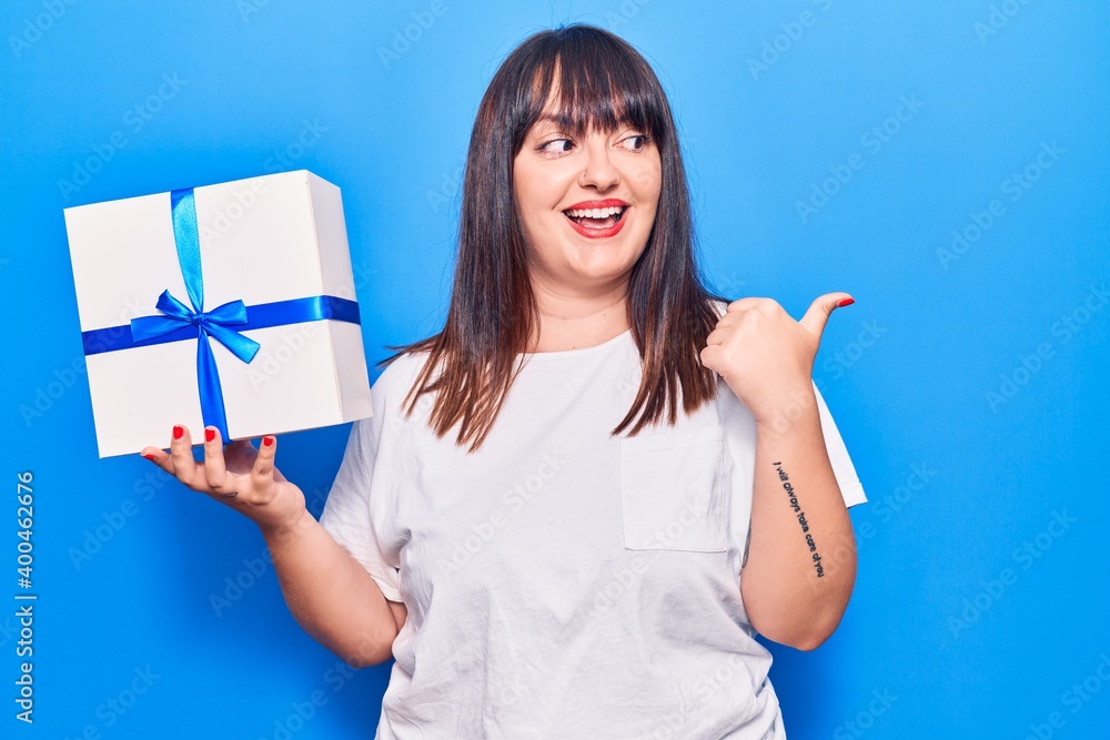 Young plus size woman holding gift pointing thumb up to the side smiling happy with open mouth Young plus size woman holding gift pointing thumb up to the side smiling happy with open mouth