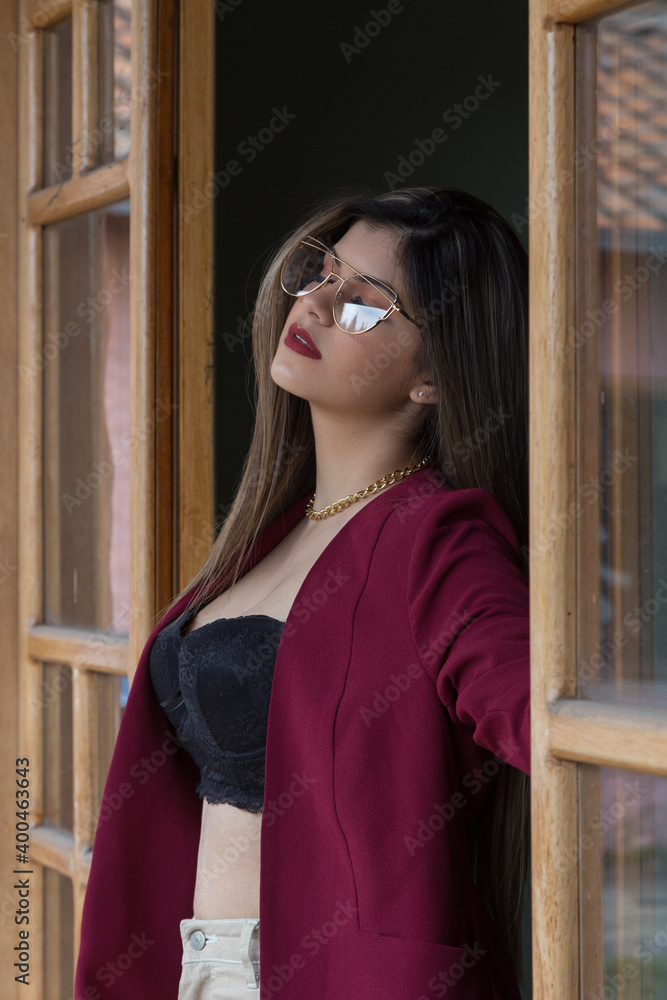 beautiful woman sitting in burgundy jacket and glasses at the window of a hut, dark background beautiful woman sitting in burgundy jacket and glasses at the window of a hut, dark background