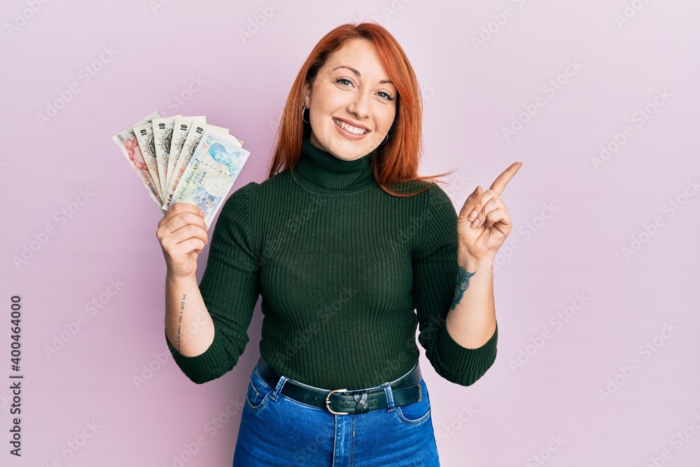Beautiful redhead woman holding united kingdom pounds smiling happy pointing with hand and finger to the side Beautiful redhead woman holding united kingdom pounds smiling happy pointing with hand and finger to the side
