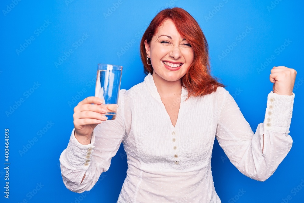 Young beautiful redhead woman drinking glass of water over isolated blue background screaming proud, celebrating victory and success very excited with raised arm Young beautiful redhead woman drinking glass of water over isolated blue background screaming proud, celebrating victory and success very excited with raised arm