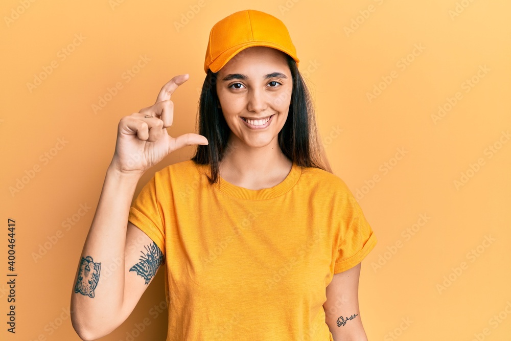 Young hispanic woman wearing delivery uniform and cap smiling and confident gesturing with hand doing small size sign with fingers looking and the camera. measure concept. Young hispanic woman wearing delivery uniform and cap smiling and confident gesturing with hand doing small size sign with fingers looking and the camera. measure concept.