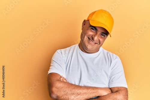 Mature middle east man with mustache wearing casual white tshirt and yellow cap happy face smiling with crossed arms looking at the camera. positive person.