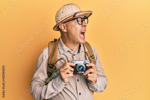 Middle age bald man wearing explorer hat and vintage camera angry and mad screaming frustrated and furious, shouting with anger. rage and aggressive concept.
