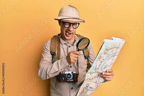 Middle age bald man wearing explorer hat holding magnifying glass on a map winking looking at the camera with sexy expression, cheerful and happy face.