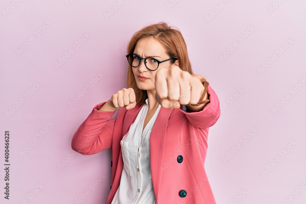 Young caucasian woman wearing business style and glasses punching fist to fight, aggressive and angry attack, threat and violence