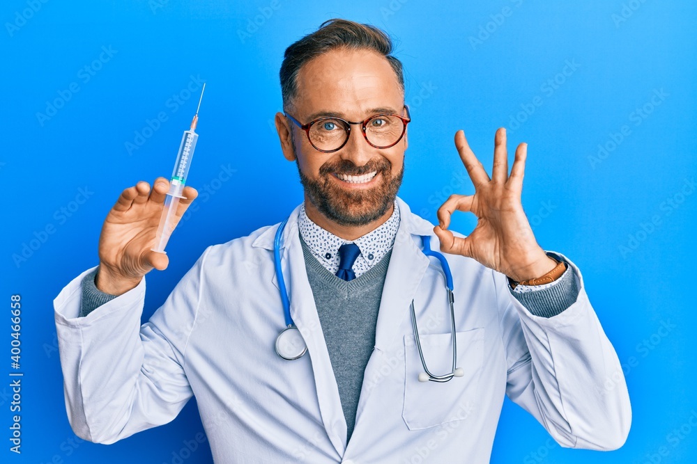 Handsome middle age man wearing doctor uniform holding syringe doing ok sign with fingers, smiling friendly gesturing excellent symbol Handsome middle age man wearing doctor uniform holding syringe doing ok sign with fingers, smiling friendly gesturing excellent symbol