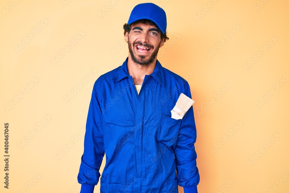 Handsome young man with curly hair and bear wearing builder jumpsuit uniform winking looking at the camera with sexy expression, cheerful and happy face.