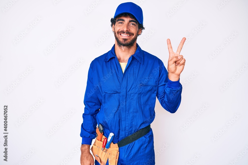 Handsome young man with curly hair and bear weaing handyman uniform showing and pointing up with fingers number two while smiling confident and happy. Handsome young man with curly hair and bear weaing handyman uniform showing and pointing up with fingers number two while smiling confident and happy.