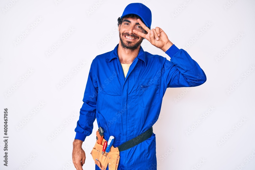 Handsome young man with curly hair and bear weaing handyman uniform doing peace symbol with fingers over face, smiling cheerful showing victory Handsome young man with curly hair and bear weaing handyman uniform doing peace symbol with fingers over face, smiling cheerful showing victory