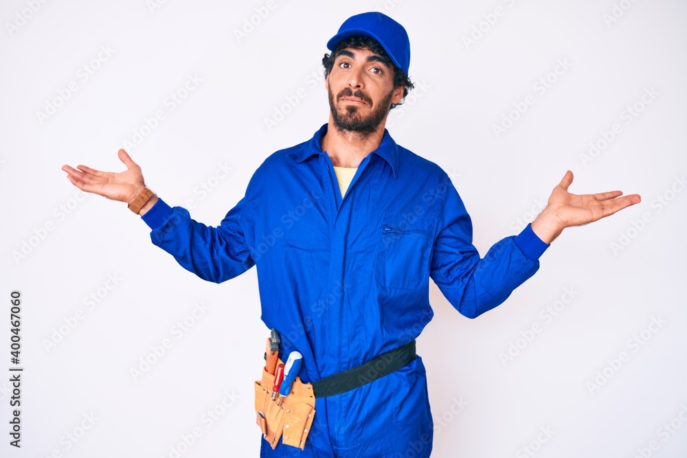 Handsome young man with curly hair and bear weaing handyman uniform clueless and confused expression with arms and hands raised. doubt concept.
