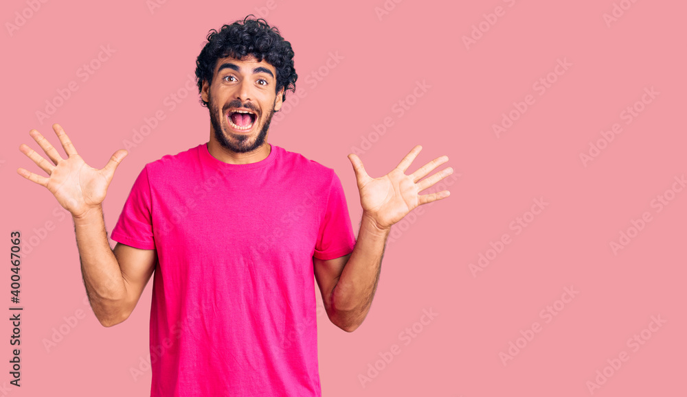 Handsome young man with curly hair and bear wearing casual pink tshirt celebrating crazy and amazed for success with arms raised and open eyes screaming excited. winner concept Handsome young man with curly hair and bear wearing casual pink tshirt celebrating crazy and amazed for success with arms raised and open eyes screaming excited. winner concept