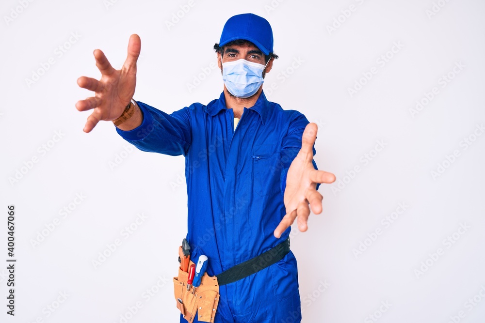 Handsome young man with curly hair and bear wearing handyman uniform and covid-19 safety mask looking at the camera smiling with open arms for hug. cheerful expression embracing happiness. Handsome young man with curly hair and bear wearing handyman uniform and covid-19 safety mask looking at the camera smiling with open arms for hug. cheerful expression embracing happiness.