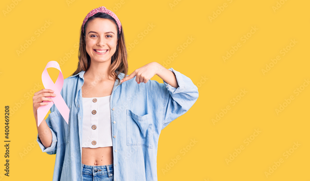 Young beautiful girl holding pink cancer ribbon pointing finger to one self smiling happy and proud Young beautiful girl holding pink cancer ribbon pointing finger to one self smiling happy and proud