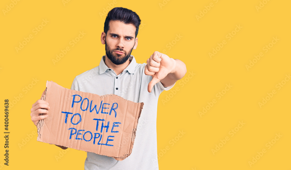Young handsome man with beard holding power to the people cardboard banner with angry face, negative sign showing dislike with thumbs down, rejection concept Young handsome man with beard holding power to the people cardboard banner with angry face, negative sign showing dislike with thumbs down, rejection concept